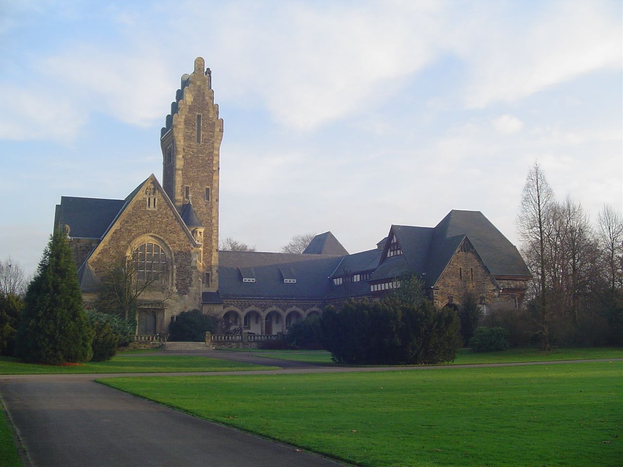 Die historische Friedhofskapelle mit hohem gestuftem Turm, großem Bogenfenster und langem Arkadengang, gesehen über Rasen und Gehölze. auf Südfriedhof Düsseldorf